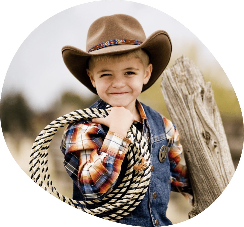 A blonde boy dressed as a cowboy carrying a rope and a log smiling to the camera
