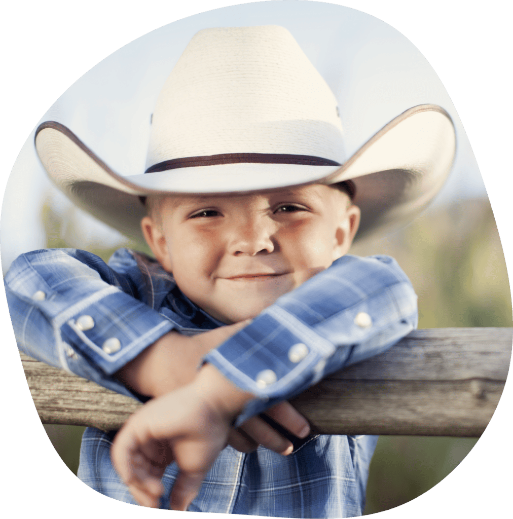 A young boy dress up as a cowboy on a ranch fence smiling to the camera